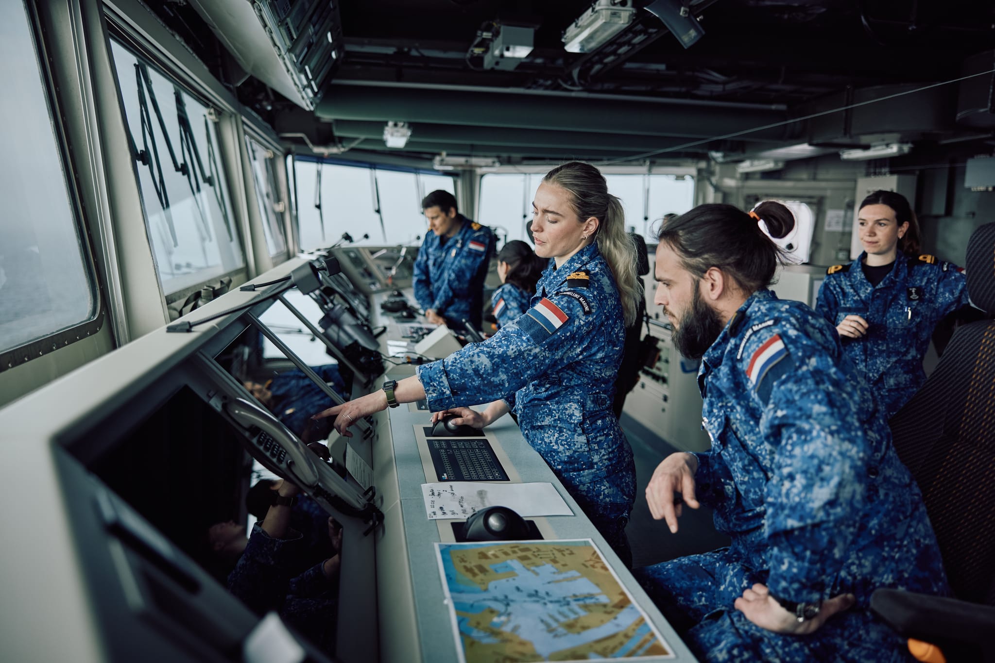 Naval officers in blue camo aboard a ship's bridge, a woman points at instruments while crew consult displays and a chart.