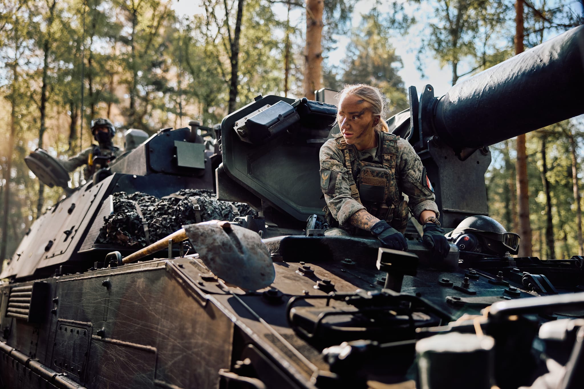 Camouflaged soldier with face paint leaning from an armored vehicle's hatch in a sunlit forest.