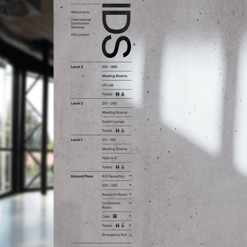 Vertical office directory sign reading "IDS" with floor listings for Levels 3–Ground (meeting rooms, UX lab, reception, toilets) in a modern lobby.