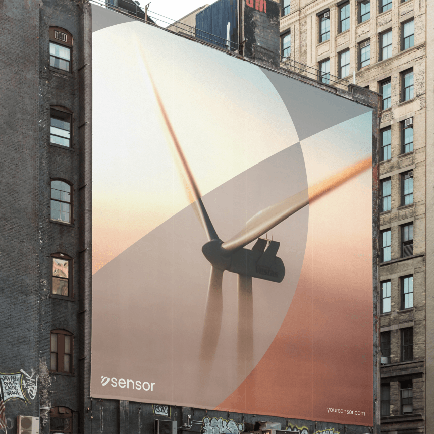 Giant rooftop billboard on an urban building displaying a close-up wind turbine against a warm gradient sky, 'sensor' logo at bottom.