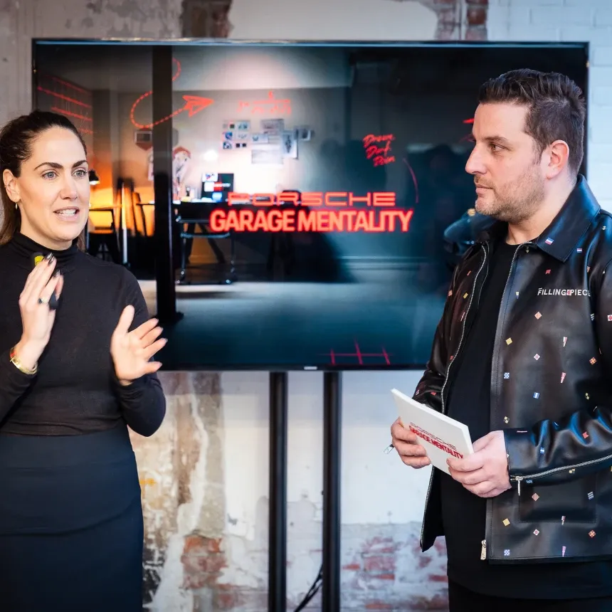 A woman and a man stand in front of a digital screen displaying the red "Porsche Garage Mentality" logo. The woman is speaking with expressive hand gestures while the man, wearing a Filling Pieces leather jacket, listens while holding a branded notebook.