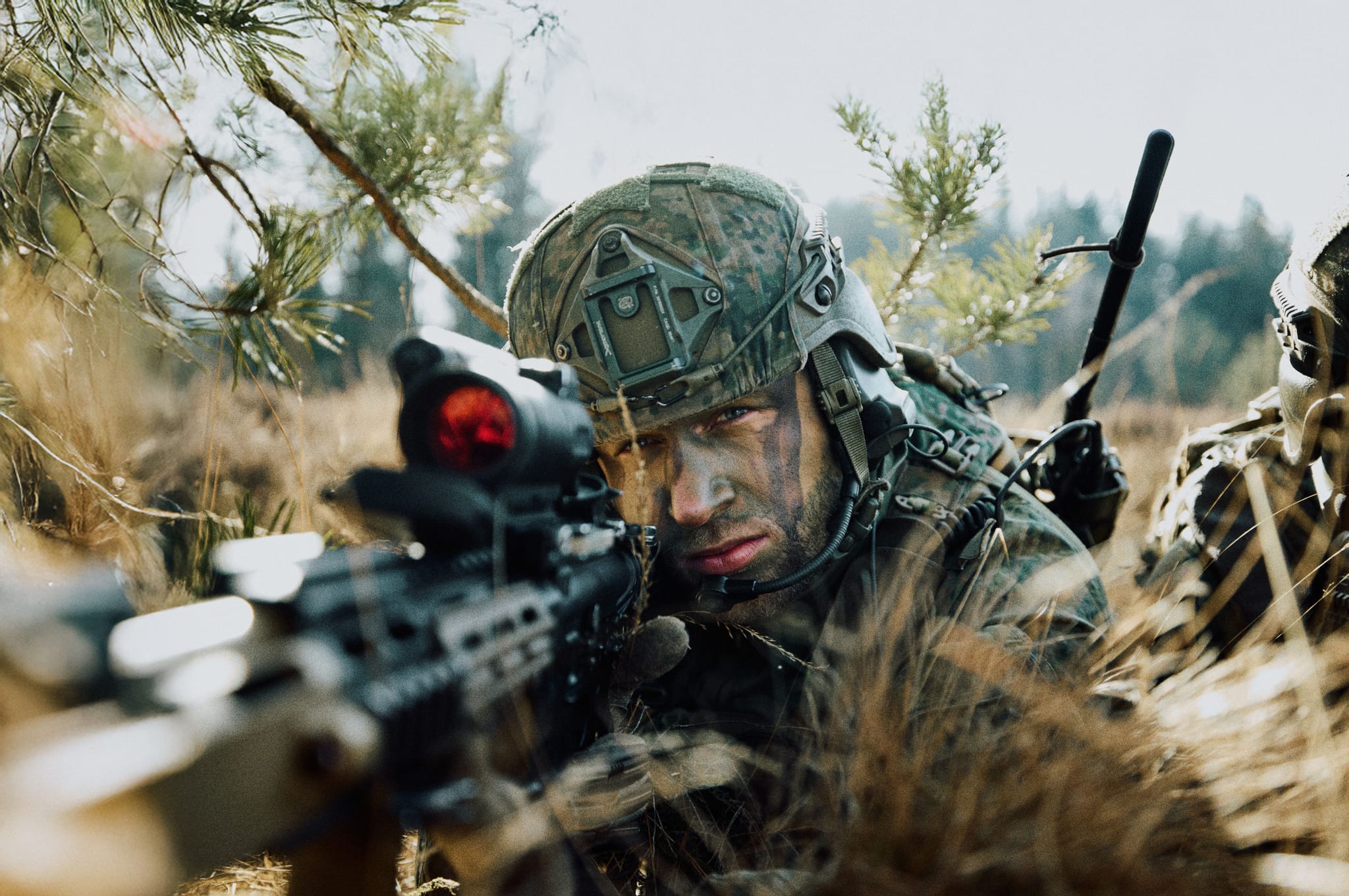 Camouflaged soldier lying in tall grass aiming a scoped rifle, wearing helmet and face paint, radio antenna visible behind him