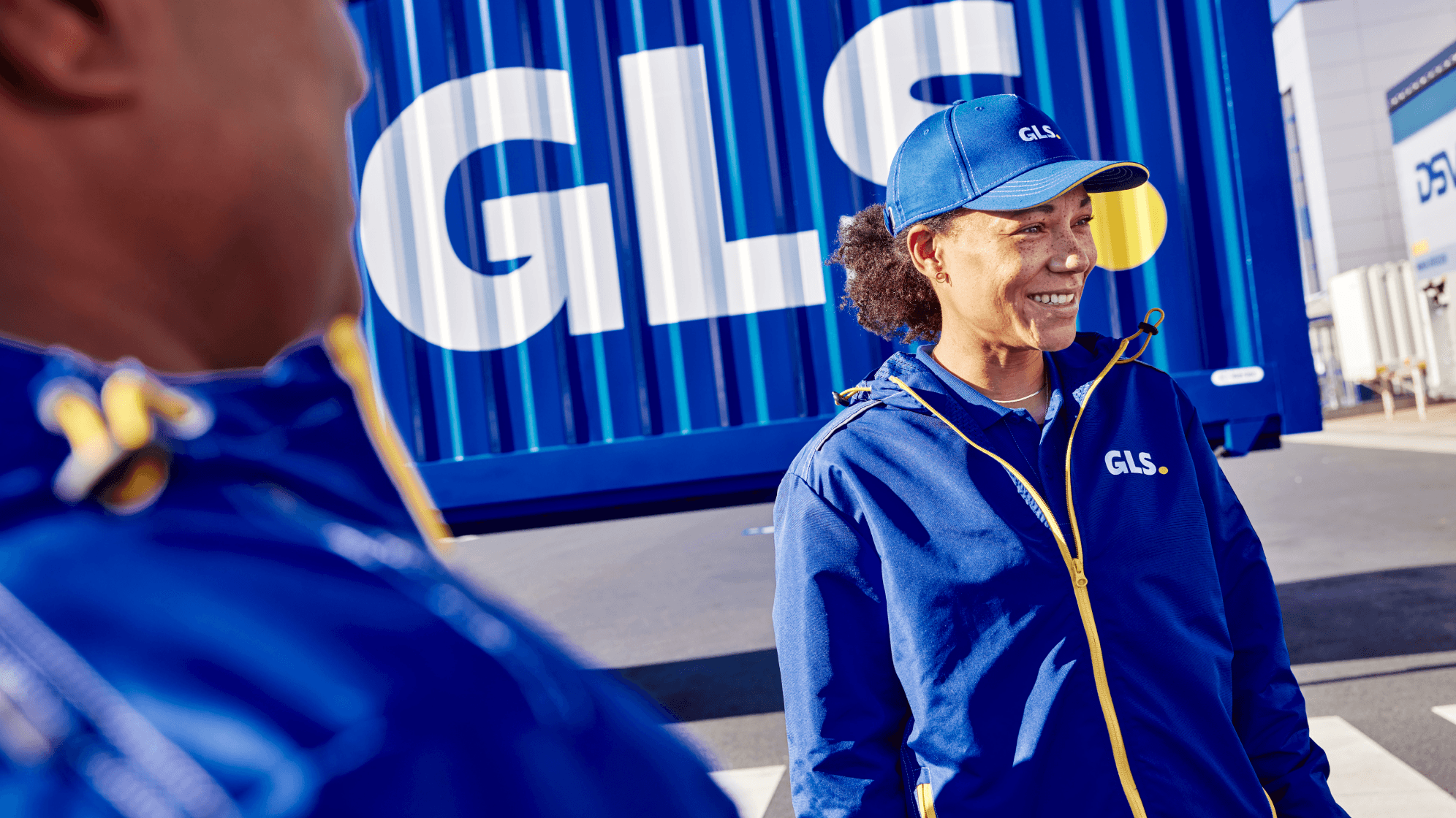 A woman in a blue GLS jacket and cap smiles while standing in front of a large, blue shipping container with the GLS logo