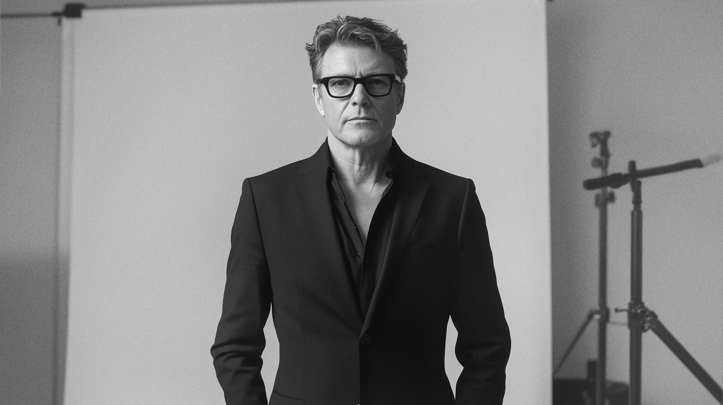 Monochrome, straight-on medium shot of an intensely serious-looking man (still the executive creative director, but now he's really feeling the weight of the market) in a dark, impeccably tailored suit, over a slightly unbuttoned dark shirt (for that 'approachable' edge). He's wearing the same signature bold glasses and staring directly into the camera lens, perhaps assessing Q4 projections. In the background, there's a seamless white backdrop and the unmistakable, casually visible silhouette of studio lighting equipment, subtly reminding us this is a professional operation, not just a casual snap. Minimalist, serious, and ready for the annual report.