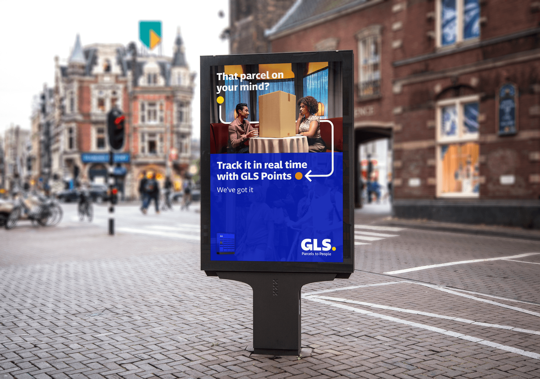 An outdoor digital advertisement on a city sidewalk features a man and a woman sitting at a table with a large cardboard box between them. The ad text reads, "That parcel on your mind? Track it in real time with GLS Points," followed by the slogan "We've got it" and the GLS logo.