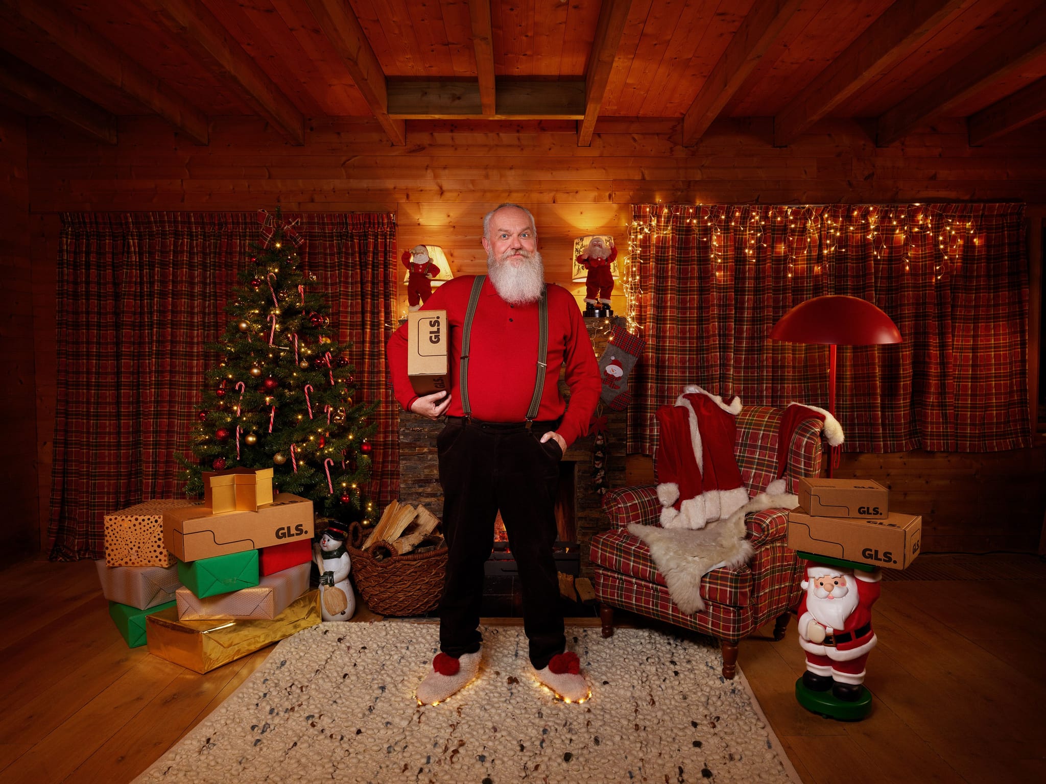 In a cozy Christmas-themed room, a man with a white beard stands holding a GLS parcel. The festive scene includes a decorated tree, glowing slippers, and several GLS-branded boxes scattered near a fireplace and armchair.