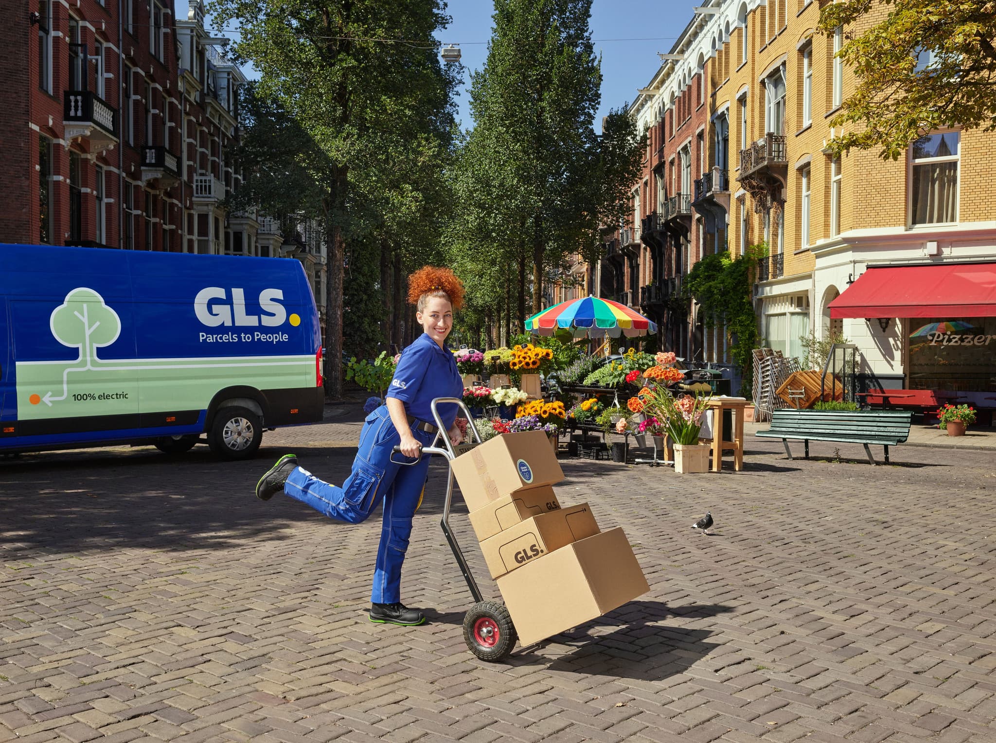 A delivery person in a blue GLS uniform poses playfully with a hand truck loaded with boxes on a cobblestone street. In the background, a blue electric GLS delivery van is parked near a colorful outdoor flower stall and classic city buildings.