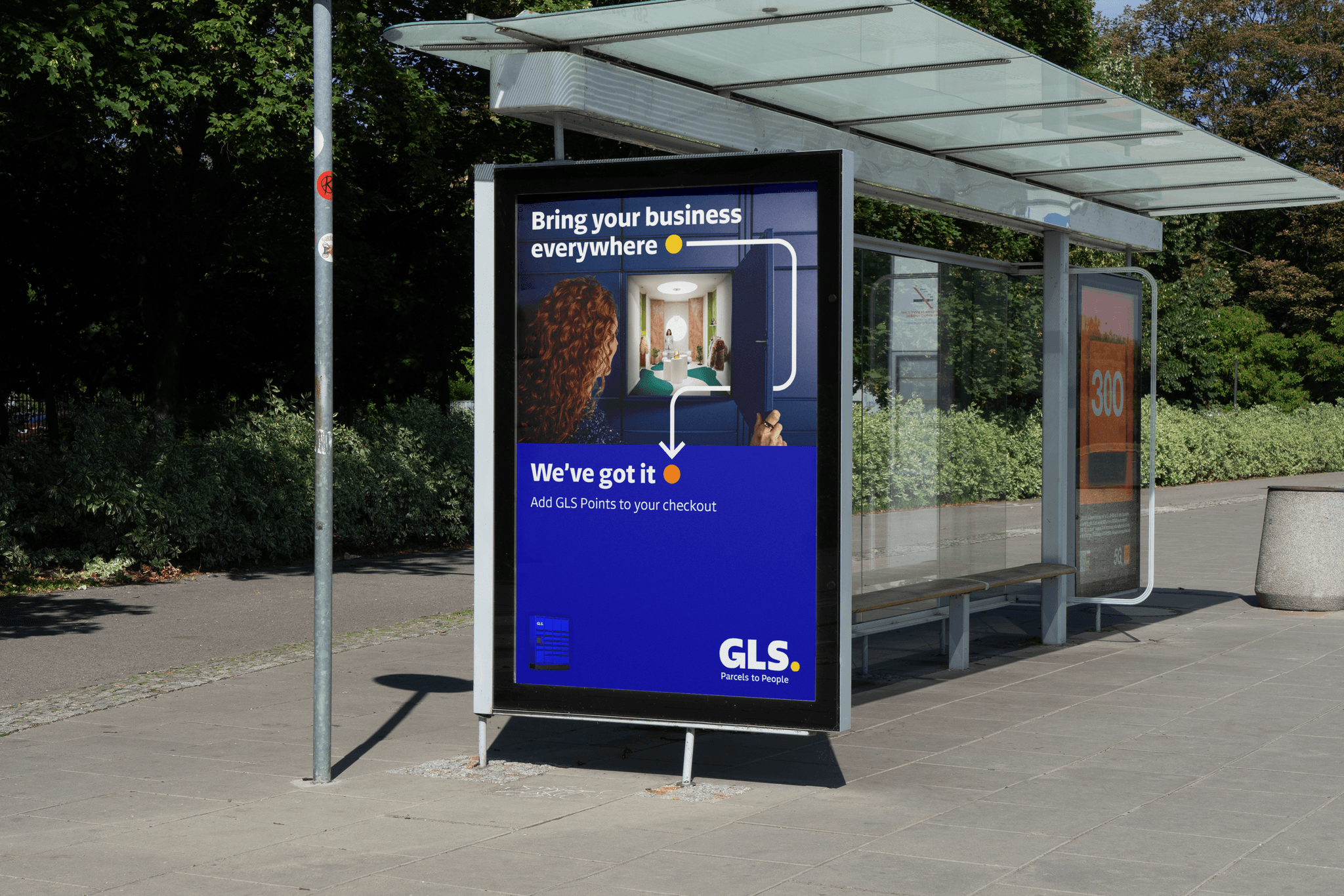 An outdoor digital advertisement at a bus stop features a woman looking into a blue parcel locker that contains a miniature boutique. The ad includes the text "Bring your business everywhere" and "We've got it," alongside the GLS logo.