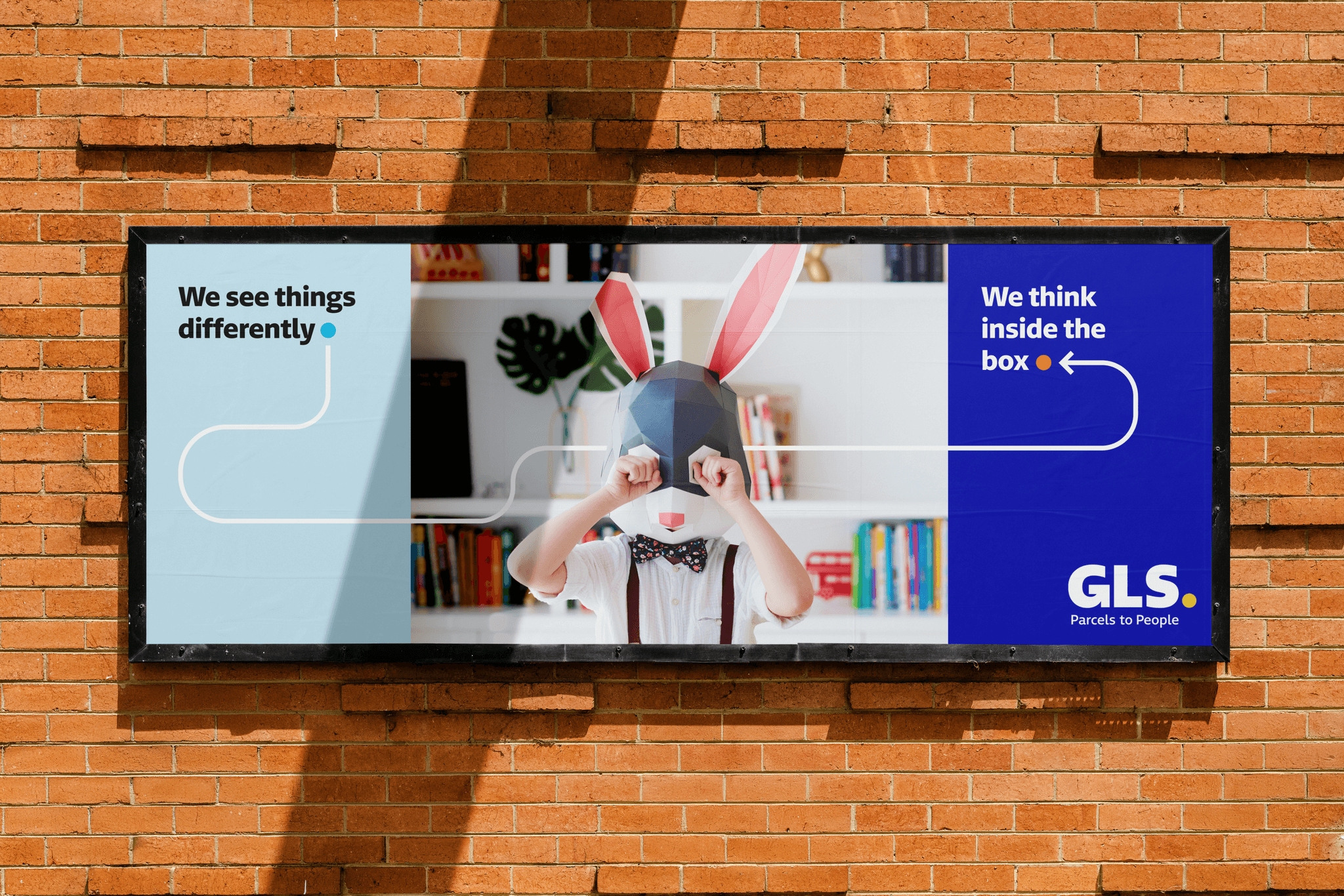 An outdoor billboard for GLS is mounted on a brick wall, featuring a child wearing a geometric rabbit mask. The ad includes the text "We see things differently" on the left and "We think inside the box" on the right, with the GLS logo in the bottom corner.