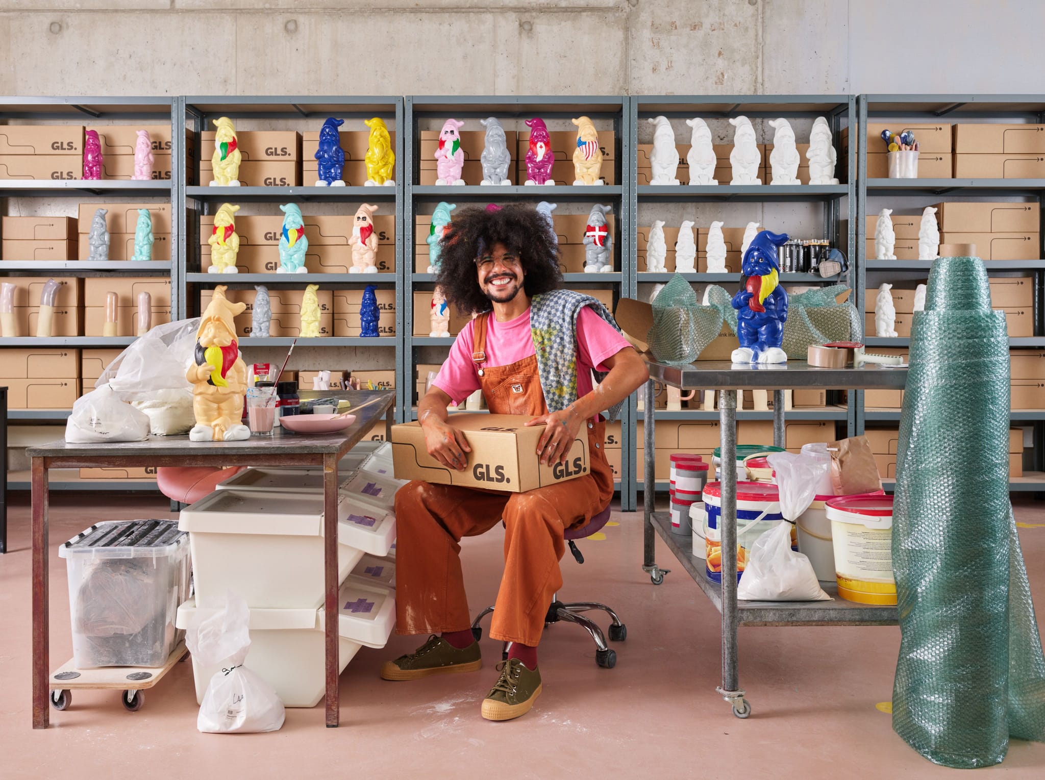 A man sits in a studio holding a GLS-branded shipping box, surrounded by shelves and tables filled with colorful garden gnomes in various stages of being painted.