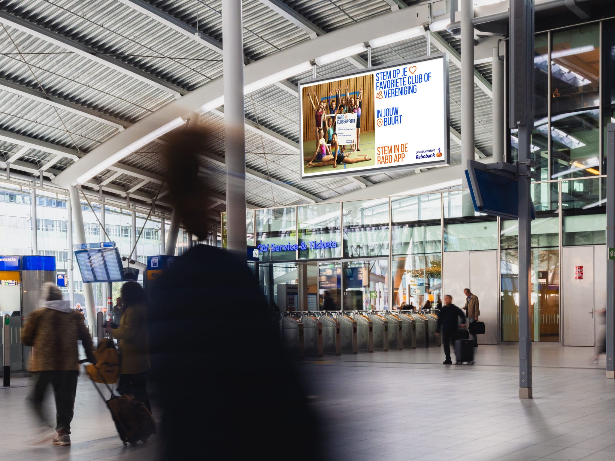 Interior of modern train station concourse with ticket barriers, digital billboard of a Rabobank ad, and blurred travelers pulling luggage.