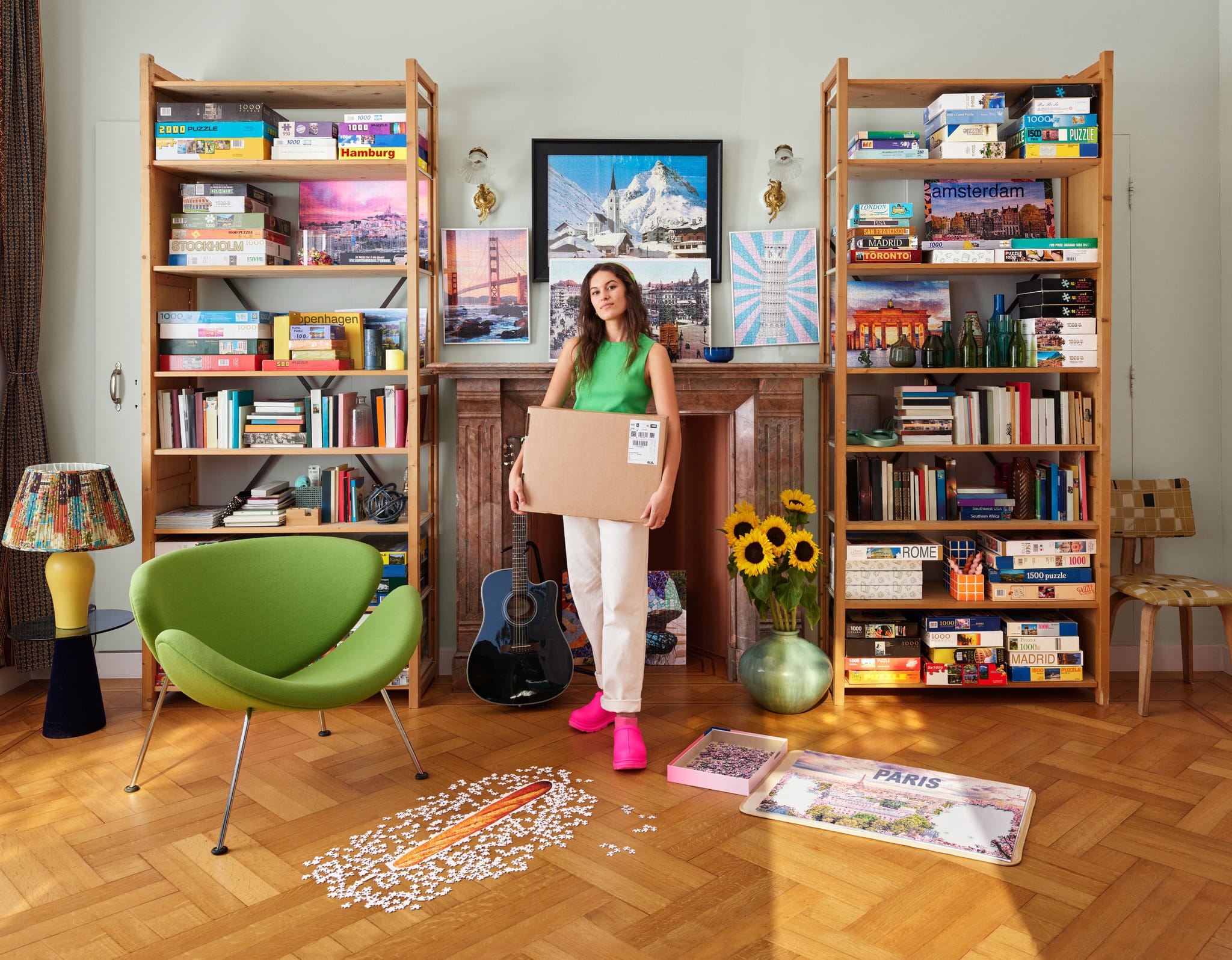 A woman in a green top stands in a room filled with puzzles, holding a large cardboard shipping box. Completed puzzles hang on the wall, and more are stacked on tall wooden shelves on either side of a fireplace.